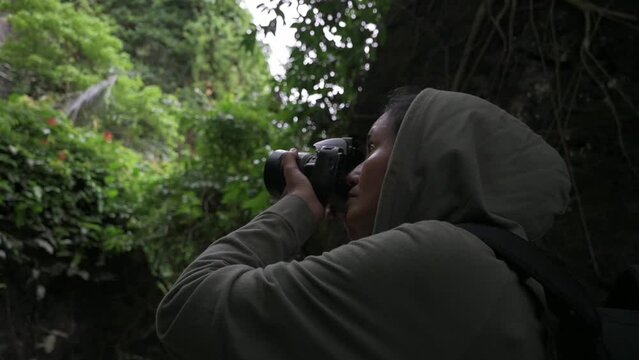 Asian woman nature photographer in hooded shirt taking pictures with camera device in tropical rainforest. Solo travel and adventure in the wild. Outdoor pursuits. Hobby and leisure.