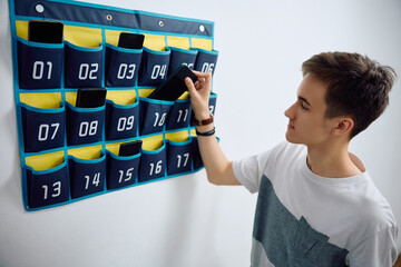 High school student leaves his smart phone in cell phone organizer on wall before entering in...