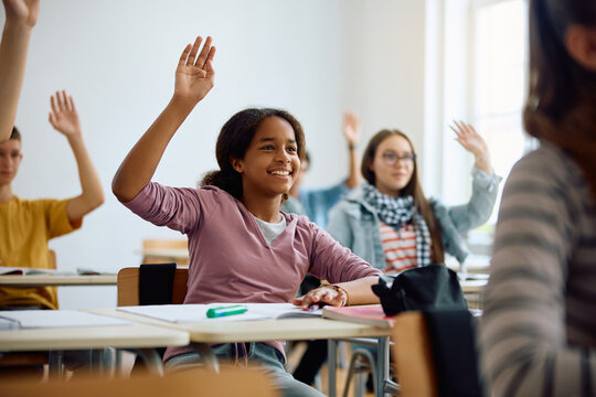 Happy Black Teenage Girl And Her Classmates Raising Hands To Answer Question During Class At High School.