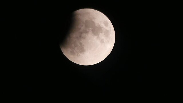 Black night sky and moving moon during a partial lunar eclipse. Moon disc with visible craters and shadow due to being partially obscured by the planet earth which was in July 2019 - time lapse.
