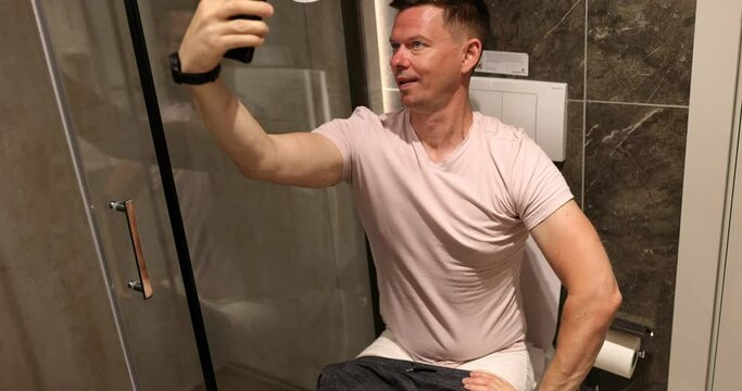 Happy man takes selfie photo sitting in bathroom on toilet. Guy grimaces and takes photos