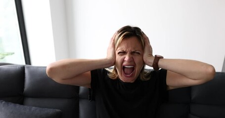 Expressive woman screaming with mouth wide open and covering ears at home on couch. Young woman emotions of anger and hysterics
