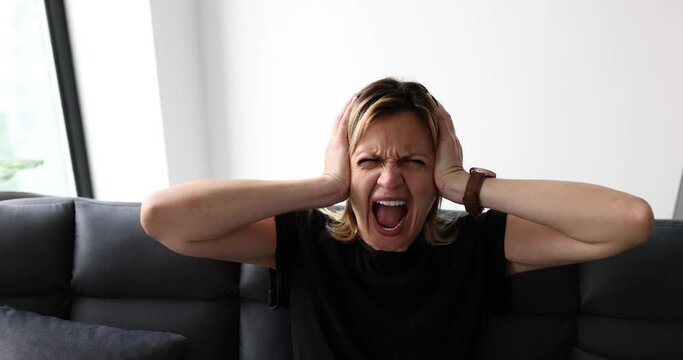 Expressive woman screaming with mouth wide open and covering ears at home on couch. Young woman emotions of anger and hysterics