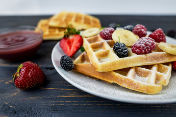 Belgian waffles with summer berries and powdered sugar in a white plate on a dark wooden background.