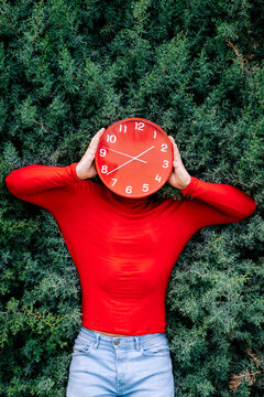 Man hiding face with red clock against green bushes