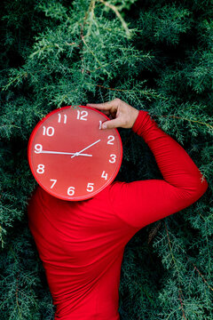 Man hiding face with red clock against green bushes