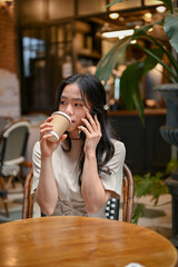 A beautiful Asian woman is sipping coffee and talking on the phone in a coffee shop.