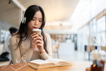 A relaxed Asian woman is sipping coffee and listening to music while reading a book in cafe