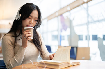 A pretty Asian woman is sipping coffee and listening to music while reading a book in a coffee shop.