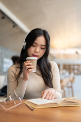 An attractive Asian woman is sipping coffee and listening to music while reading a book