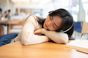 A beautiful Asian woman is listening to music on her headphones and falling asleep on a table