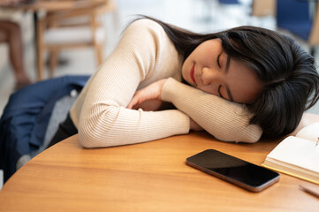 A beautiful young Asian woman is falling asleep on a table in a library or coffee shop.