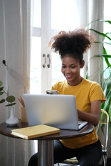 Smiling African American woman sitting in cozy home working remotely, surfing internet on laptop