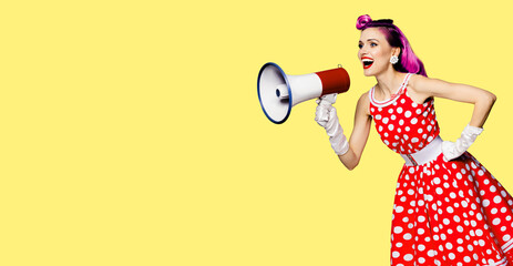 Red purple haired woman holding megaphone, shout advertising something. Girl in pin up style dress in polka dot. Yellow colour background with mock up. Female model in retro fashion vintage photo.