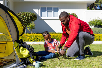Happy african american son and father pitching tent together in sunny garden