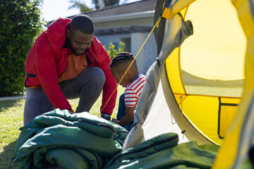 Happy african american son and father pitching tent together in sunny garden