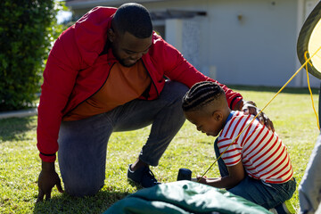 Happy african american son and father pitching tent together in sunny garden