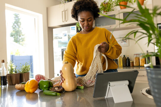 Plus size african american woman unpacking groceries and using tablet in kitchen