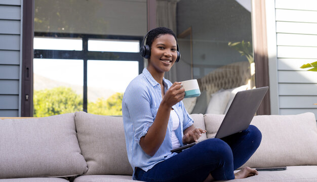 Portrait of happy african american woman wearing headphones sitting on sofa using laptop on terrace