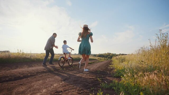 Mom dad teaching son ride bike together in village on summer vacation. Parents running near child boy father holding bicycle clapping hands supporting kid. Family weekend on nature, outdoors activity.