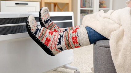 Closeup of woman wearing warm socks sitting at electric heater in living room. Concept of energy crisis, high bills, economy and saving money on monthly utility payments.