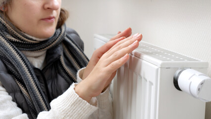 Closeup of woman in scarf warming hands at heating radiator in house. Concept of energy crisis, high bills, broken heating system, economy and saving money on monthly utility payments