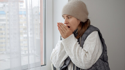 Young woman in hat and sweater freezing at home looking out of the window. Concept of energy crisis, high bills, broken heating system, economy and saving money on monthly utility payments.
