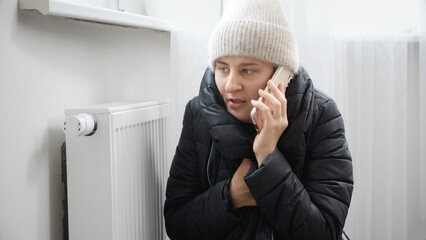 Portrait of stressed brunette woman in winter coat and woolen hat calling service to repair broken heater radiator - obrazy, fototapety, plakaty