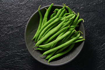 Many green pods of raw asparagus beans in a bowl on a black stone background. Top view.
