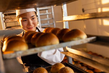 Happy asian female baker in bakery kitchen wearing apron and holding baking tray with rolls
