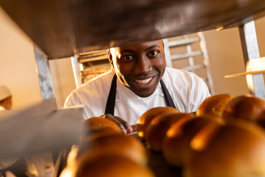 Portrait of happy african american male baker in bakery kitchen with fresh rolls