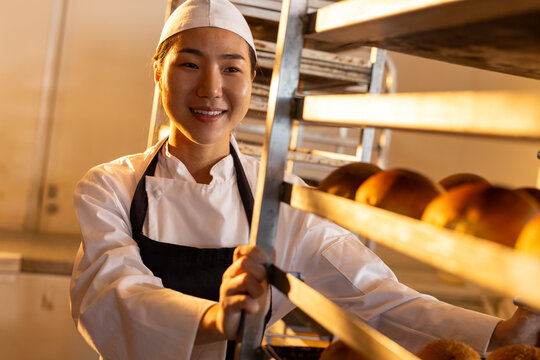 Happy asian female baker in bakery kitchen wearing apron with baking trays with rolls
