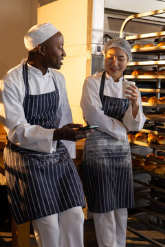 Happy diverse bakers wearing aprons in bakery kitchen and using smartphone with takeaway coffee