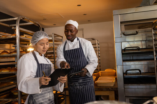 Focused diverse bakers wearing aprons in bakery kitchen, holding clipboard and using tablet