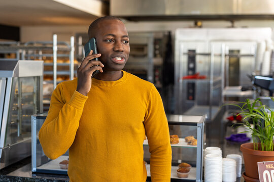 Happy African American Male Bakery Worker Wearing Yellow Sweatshirt And Talking On Smartphone