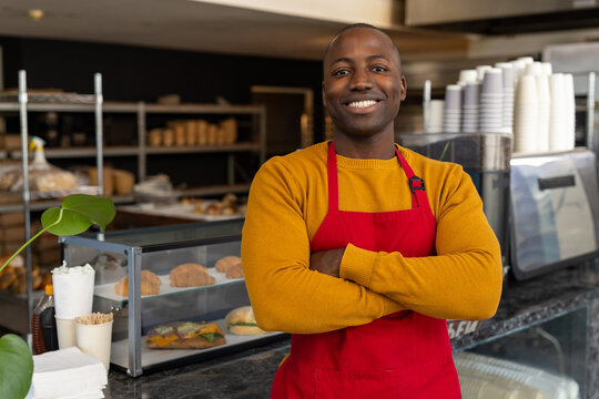 Portrait of happy african american male bakery worker wearing red apron with arms crossed
