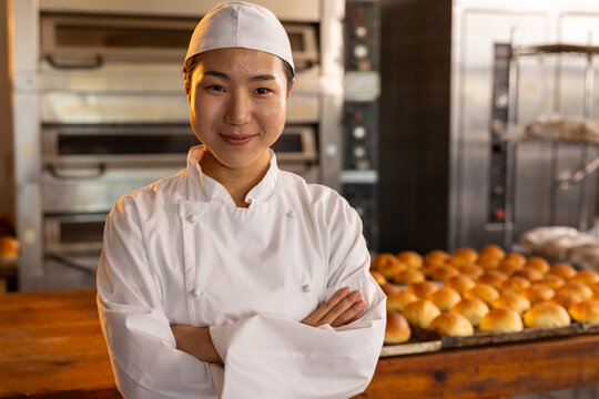 Portrait of happy asian female baker with arms crossed in bakery kitchen over fresh rolls