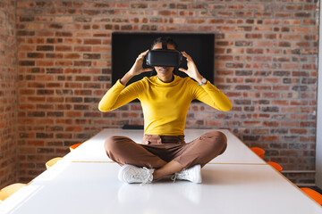 African american casual businesswoman sitting on table and using vr headset in office