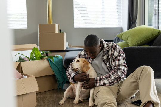 Happy african american man moving house, sitting on floor, embracing his pet dog at home,