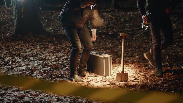 Close-up view of black case and shovel in the ground in autumn forest with fallen leaves. Forensic scientists working in nighttime crime scene. Detective thriller vibe