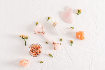 A set of cosmetics and a quartz roller massager with gua sha on a white textured background among flowers and lumps of bath salt. Top view. Flat lay.