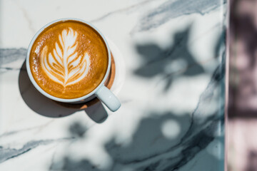 Top view of cup of tasty cappuccino with latte art on marble table background with shadows at sunny day outdoor.