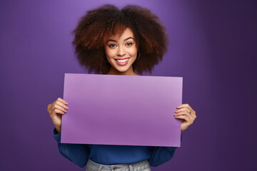 Cheerful african american woman holding a rectangular template for text in her hands on a purple plain background. Mocap, banner, copy space.