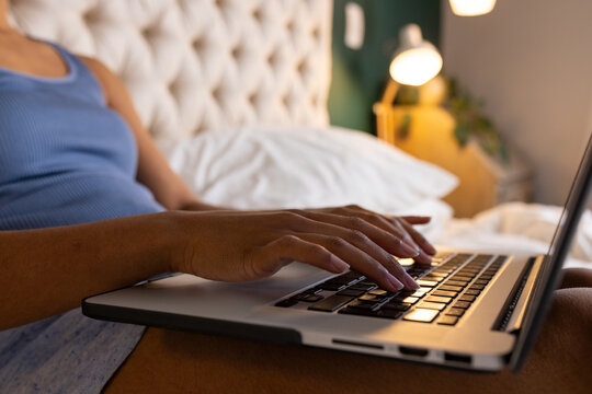 Midsection of biracial woman sitting on bed and using laptop in bedroom