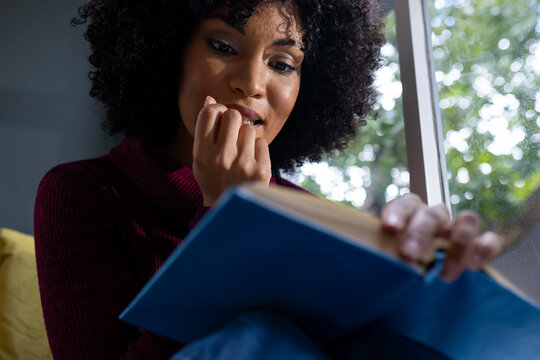 Thoughtful biracial woman sitting by window and reading book at home