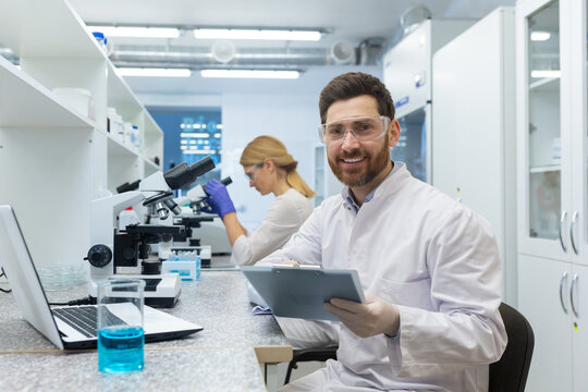 Portrait of a young male scientist looking into the camera with a smile. He is sitting in the laboratory in front of a microscope with a documents, a female colleague in the background. - Powered by Adobe