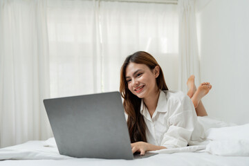 Beautiful happy asian woman lying in bed relaxed and looking at laptop screen. Girl smiling happy while browsing internet on computer, resting at home