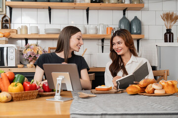 Happy two young women working cooking while her is reading a cookbook. Two young diverse lesbian women spending time together. LGBT and gender identity concept