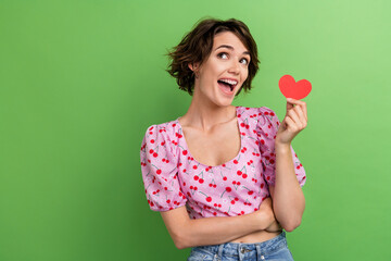 Portrait of impressed girlish ecstatic girl wear stylish blouse hold red heart staring at empty space isolated on green color background
