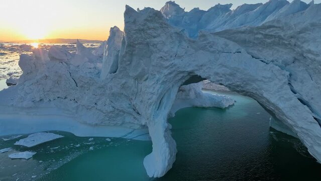 Aerial view of setting sun breaks through the ledges of iceberg. Iceberg with ice cave at sunset. Glaciers melting, global warming, arctic nature landscape